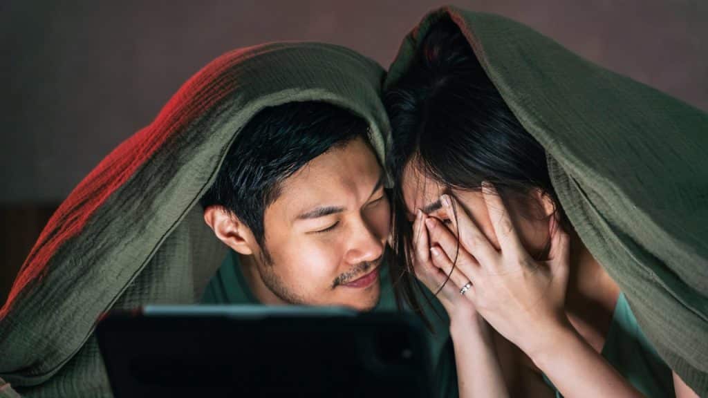 A woman hiding her face with her hands while watching a movie with her husband in a blanket.