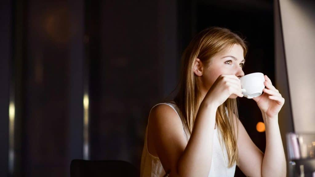 A woman drinking from a white cup while sitting indoors.