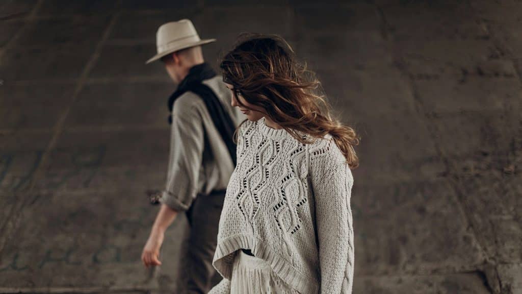 A woman in a light cable-knit sweater is standing, with a man walking away in the background.