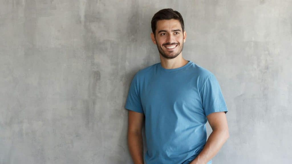 Smiling man in a blue t-shirt with his hands in his pockets, standing by a gray wall.