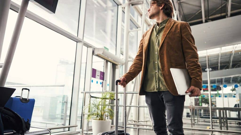 A man in a brown jacket stands in an airport terminal, holding a laptop and a suitcase handle.