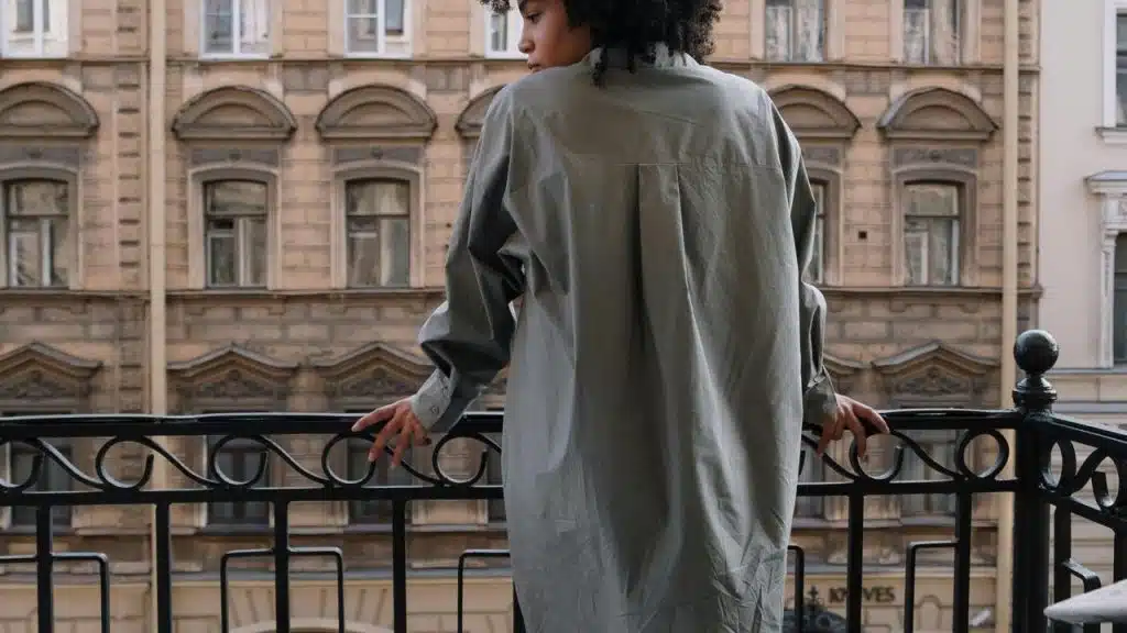 A person with curly hair in an oversized shirt standing on a balcony.