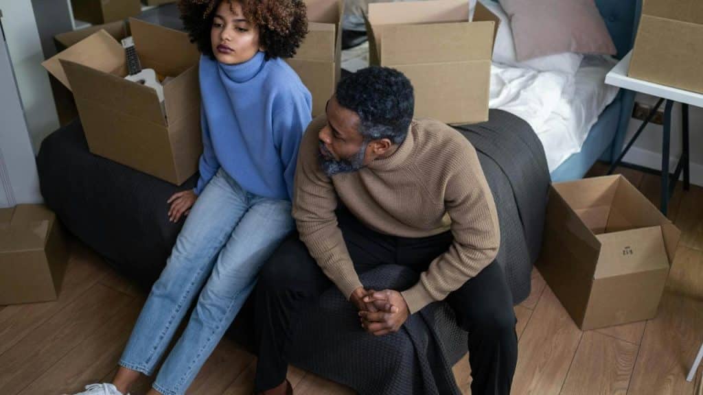 A couple sitting on a bed surrounded by moving boxes, both looking upset and distant.