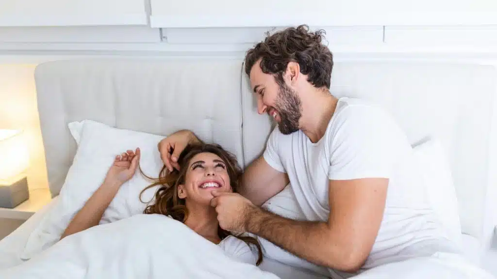 A smiling man and woman are lying in bed, looking at each other and laughing.