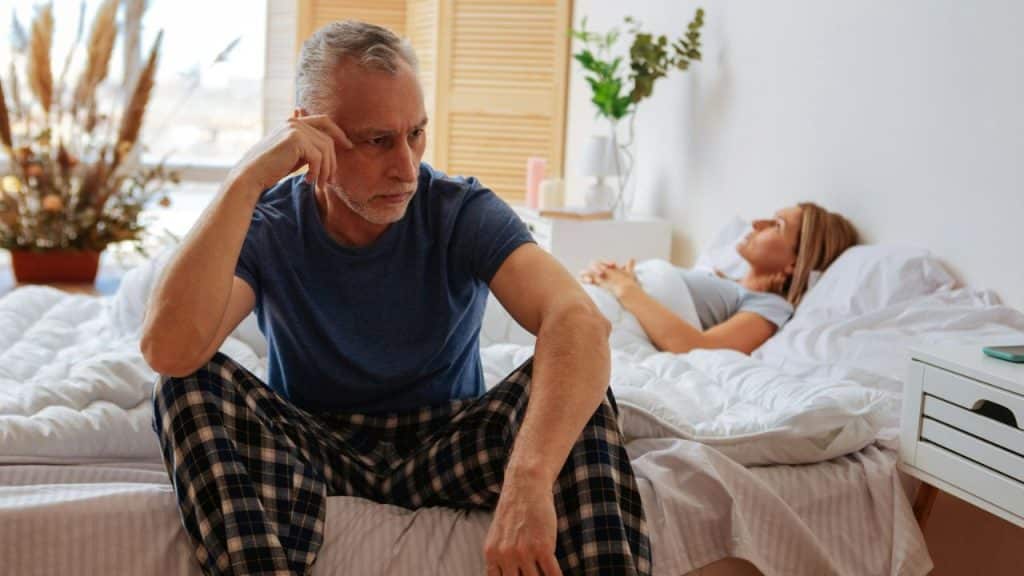 A sad-looking older man is sitting on the edge of a bed while a woman sleeps behind him.