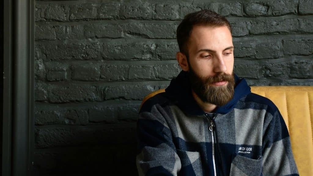 A serious man is sitting in a plaid jacket, looking down against a dark brick wall.