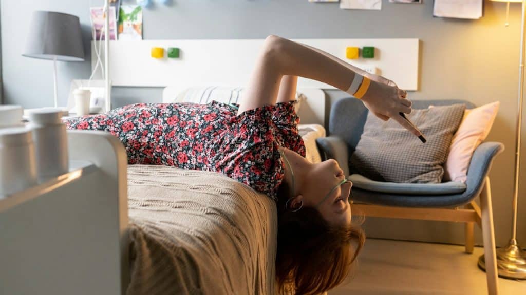 A young person in a floral dress lies upside down on a bed, looking at and holding a smartphone.