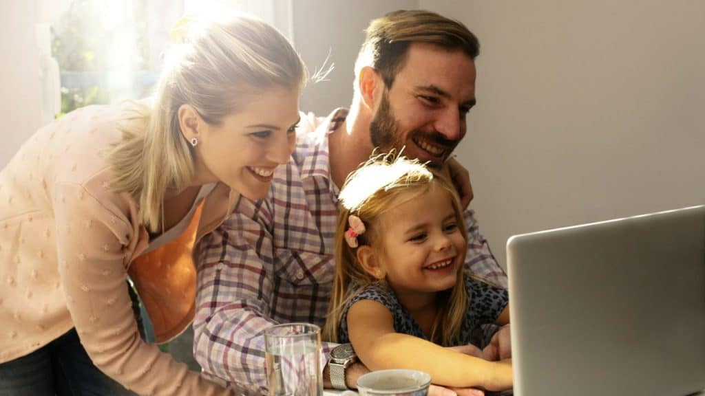 A family, including a man, woman, and girl, smiling while looking at a laptop.