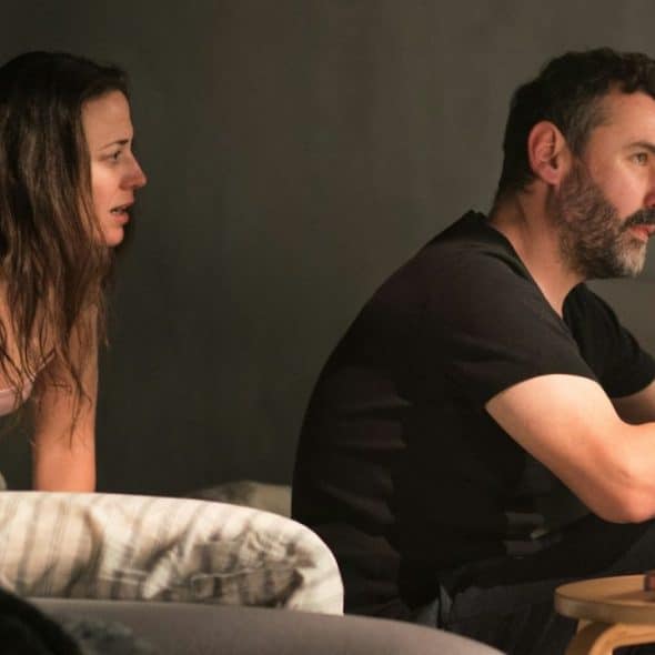 A woman in a tank top and a bearded man sit on a bed or couch, talking intensely.