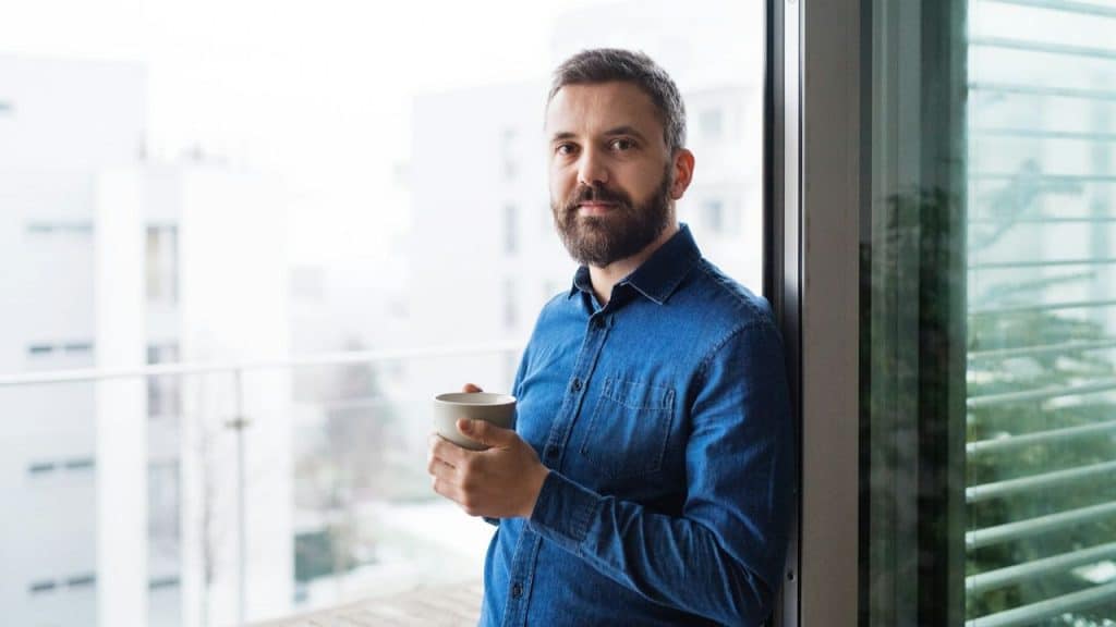 A bearded man in a blue shirt holds a cup while standing on a balcony.