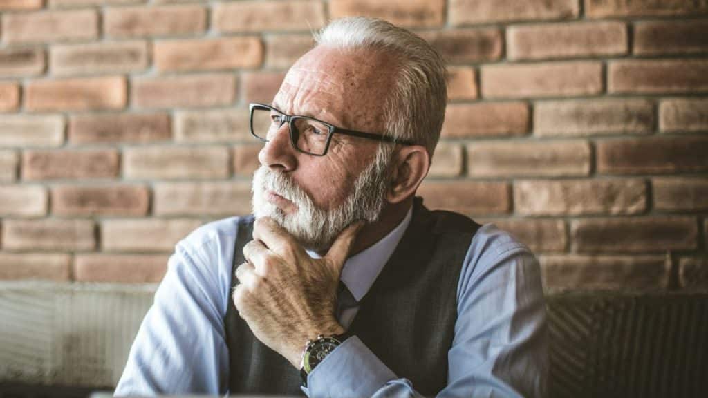 An older man with a white beard and glasses holds his chin thoughtfully, facing left.