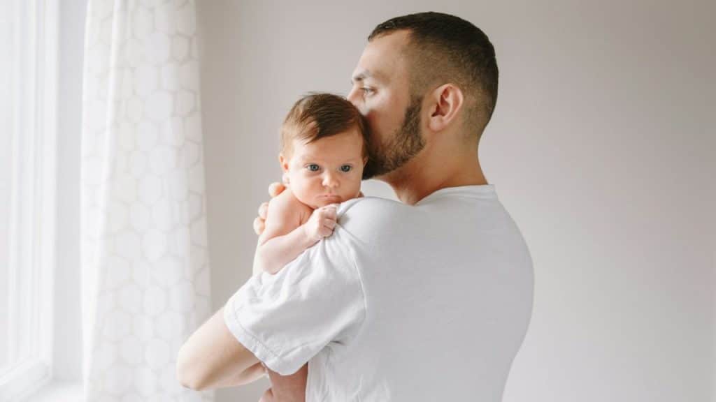 A man in a white shirt holds a newborn baby over his shoulder, kissing the baby's head.