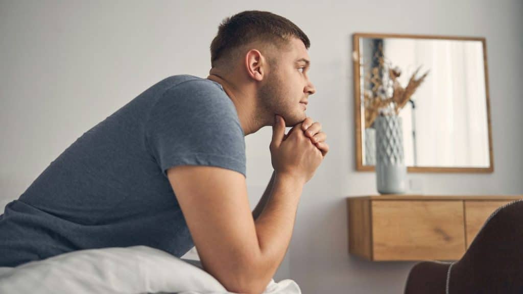 Man with a short haircut in a blue t-shirt sitting on a bed, looking off thoughtfully.