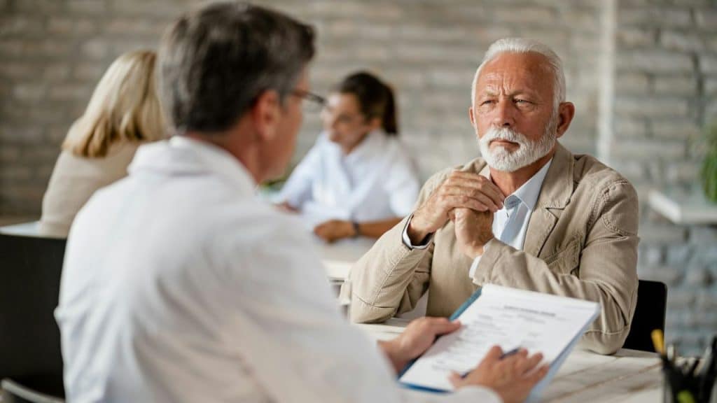 Older man with a white beard sitting across a table from a doctor, who is holding a clipboard.