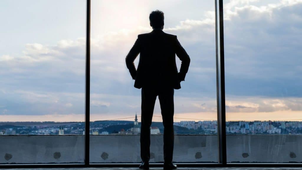A silhouetted businessman stands looking over a city from a large window.