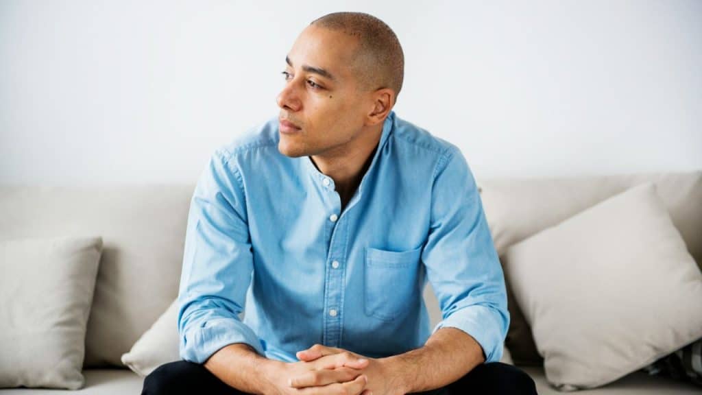 Bald man in light blue shirt sits on a beige couch, looking away.