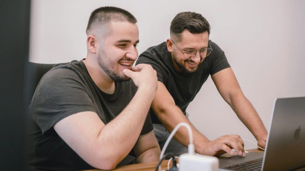 Two men smile while looking at a laptop computer on a desk in a bright room.