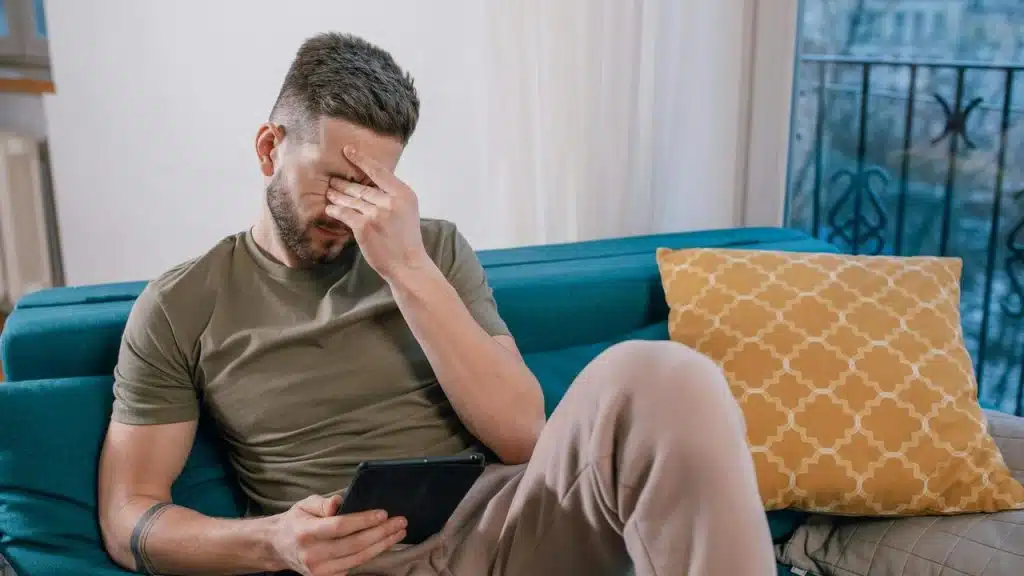 A man sitting on a couch holding a tablet with his hand covering his face.