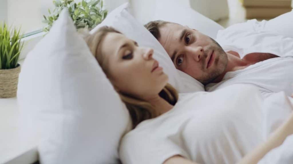 Man sitting on the bed looking conflicted while his partner sleeps