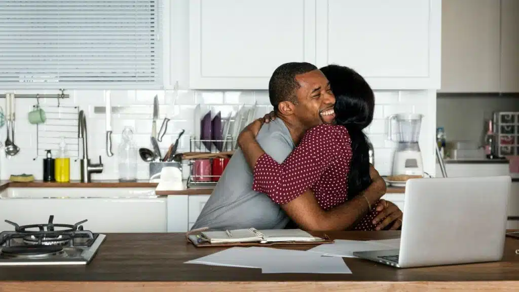 A smiling man and woman hug across a kitchen counter with a laptop and papers.