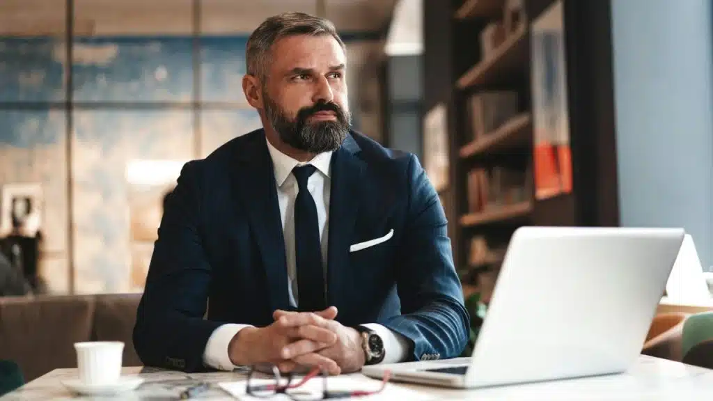 A bearded man in a navy suit sits at a table with a laptop and coffee, looking to the side.