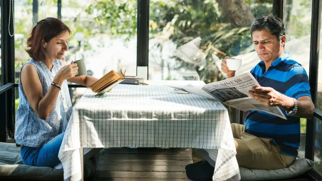 A man reads a newspaper and a woman sips a drink at a sunny outdoor table.