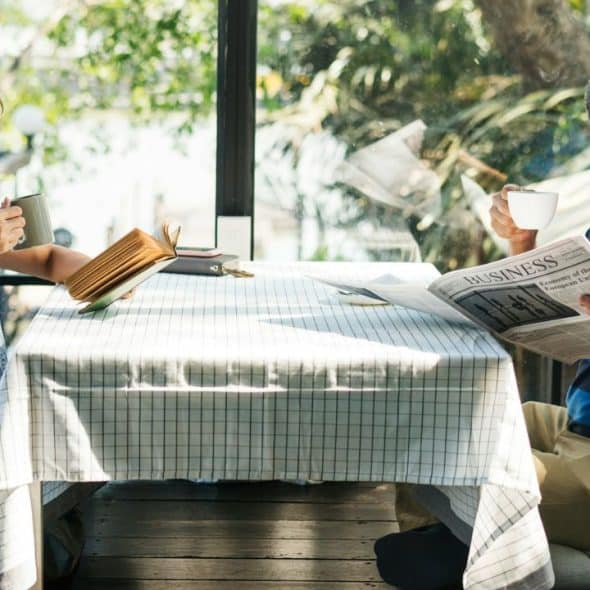 A man reads a newspaper and a woman sips a drink at a sunny outdoor table.