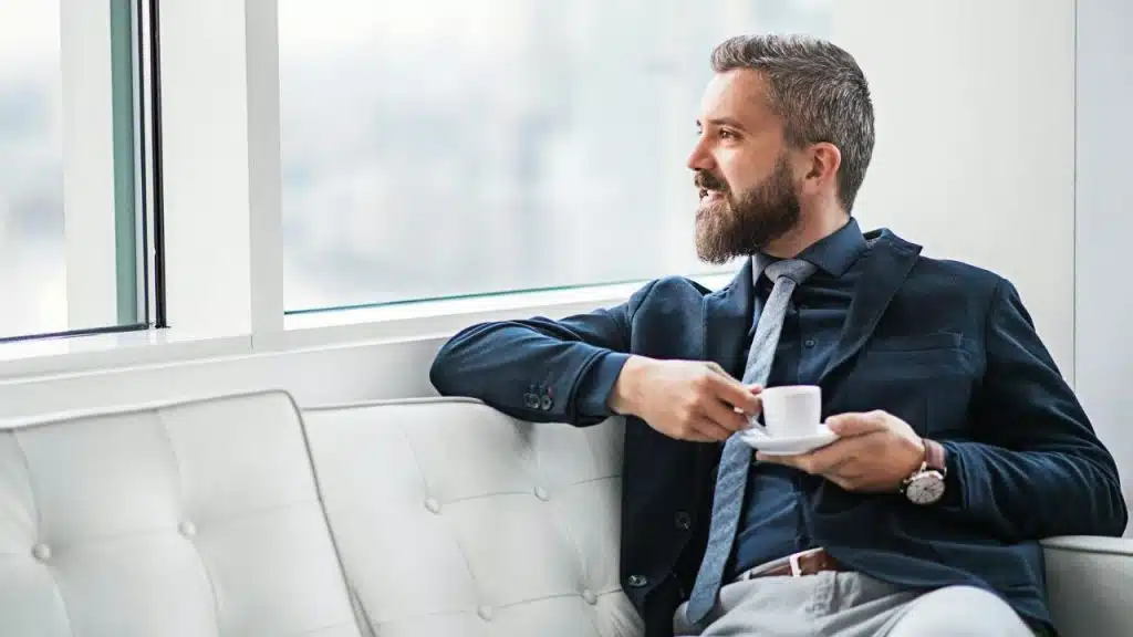A smiling, bearded businessman sits on a white couch holding a small cup of coffee.