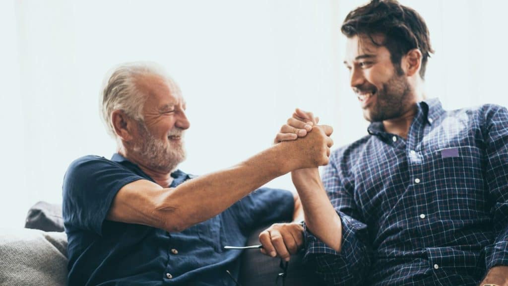 Two men, one older with a beard and one younger, happily shaking hands or clasping arms while sitting together.