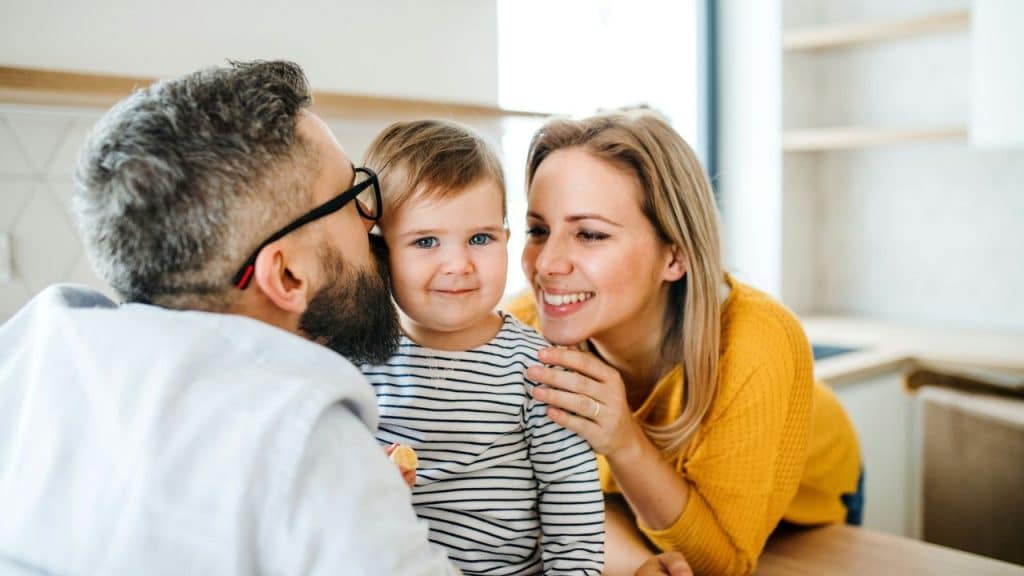 Man with a beard and glasses kisses a smiling toddler as a woman smiles beside them.