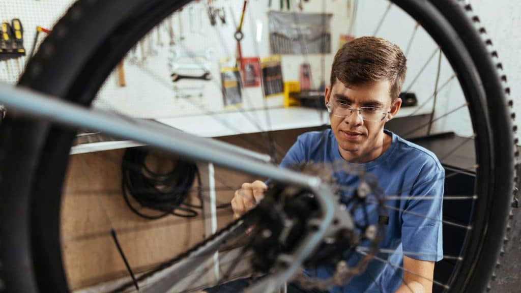 Man in glasses and a blue shirt repairing a bicycle wheel in a workshop.