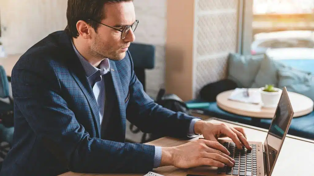 Man in glasses and a plaid blazer working seriously on a laptop in a cafe.