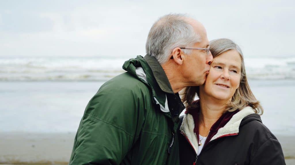 An older man kissing his wife on the cheek at the beach.