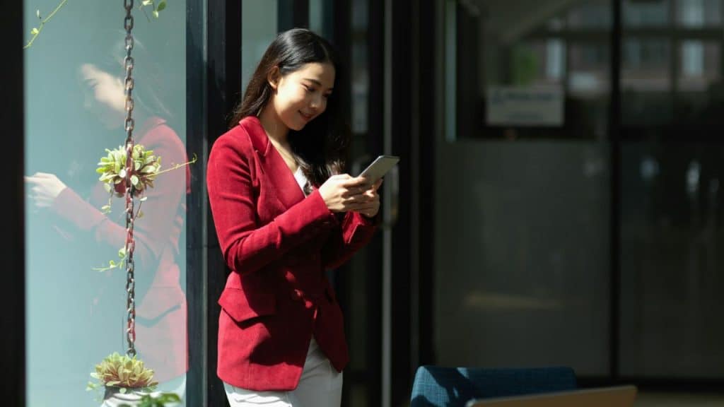 A young woman in a red blazer is standing and smiling while looking at her phone.