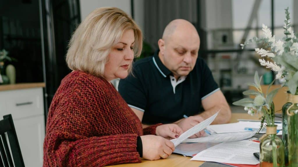 A serious middle-aged couple is sitting at a table, looking over documents in their home.