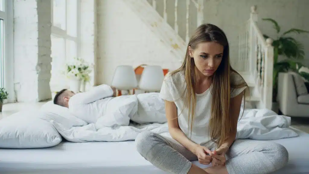 A sad woman sitting on a bed while her husband sleeps.