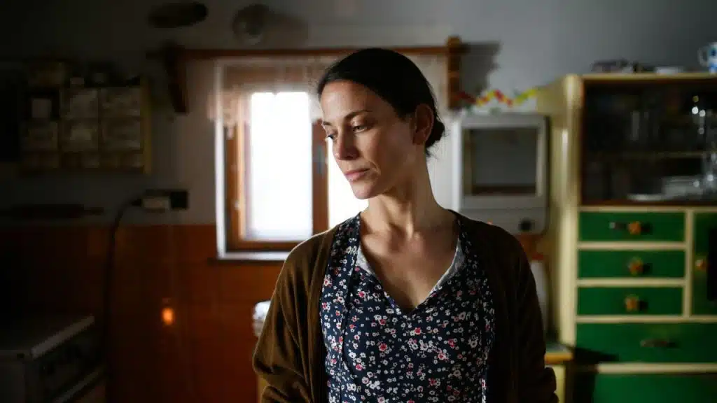 A woman in a floral top and brown cardigan stands in a dimly lit kitchen, looking down with a somber expression.
