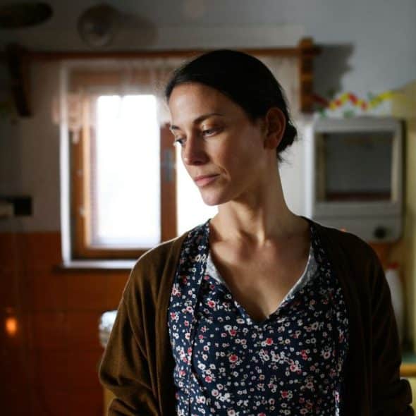 A woman in a floral top and brown cardigan stands in a dimly lit kitchen, looking down with a somber expression.