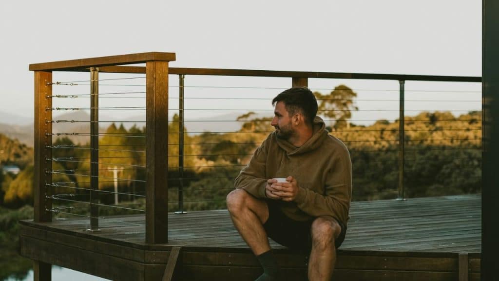 A man sits alone on a wooden deck at sunset, holding a white mug and looking out at the scenery.