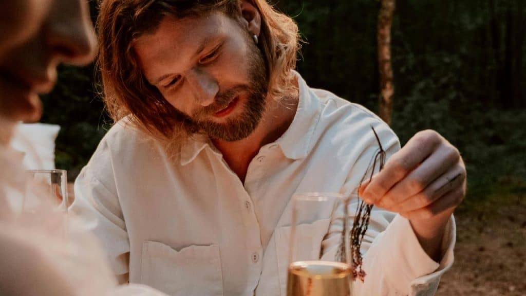 A man with long hair and a beard sitting outdoors wearing a white shirt.