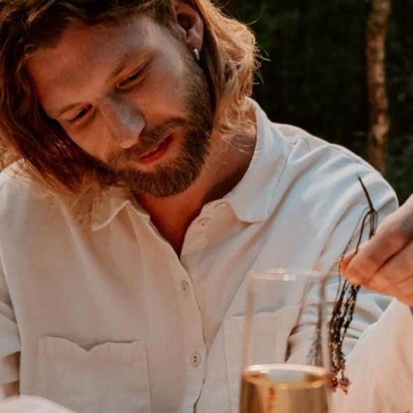 A man with long hair and a beard sitting outdoors wearing a white shirt.