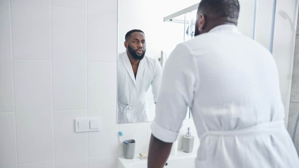 Black man in a white robe looking into a bathroom mirror.
