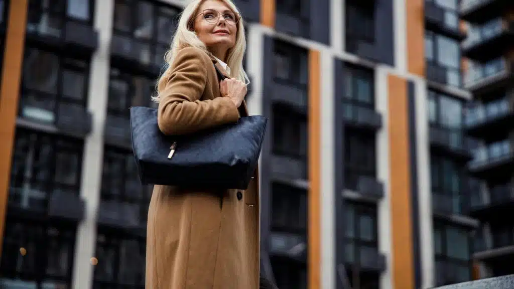 A woman in a brown coat holding a black bag while standing outside modern buildings.
