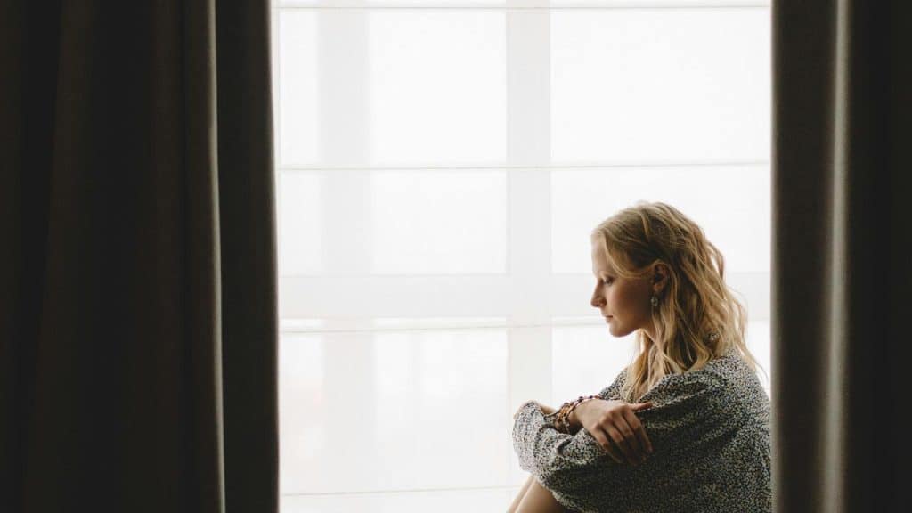 A woman sitting by a window with curtains, looking thoughtful.