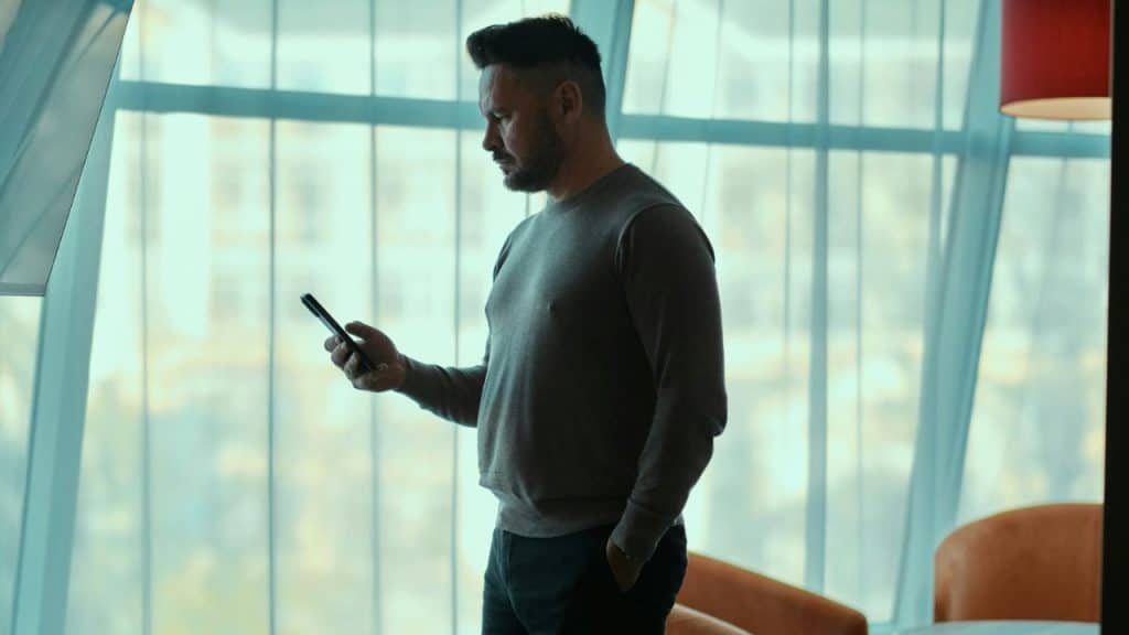 A man in a sweater is standing near a large window, looking at his smartphone.
