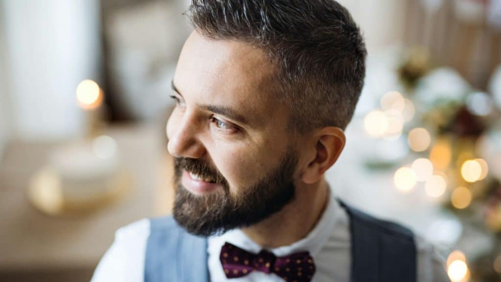 Close-up of a smiling man with a beard, bow tie, and vest looking to the side.