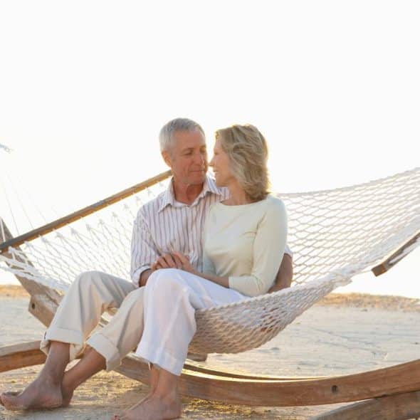 A senior couple sitting on a hammock at the beach, looking at each other.
