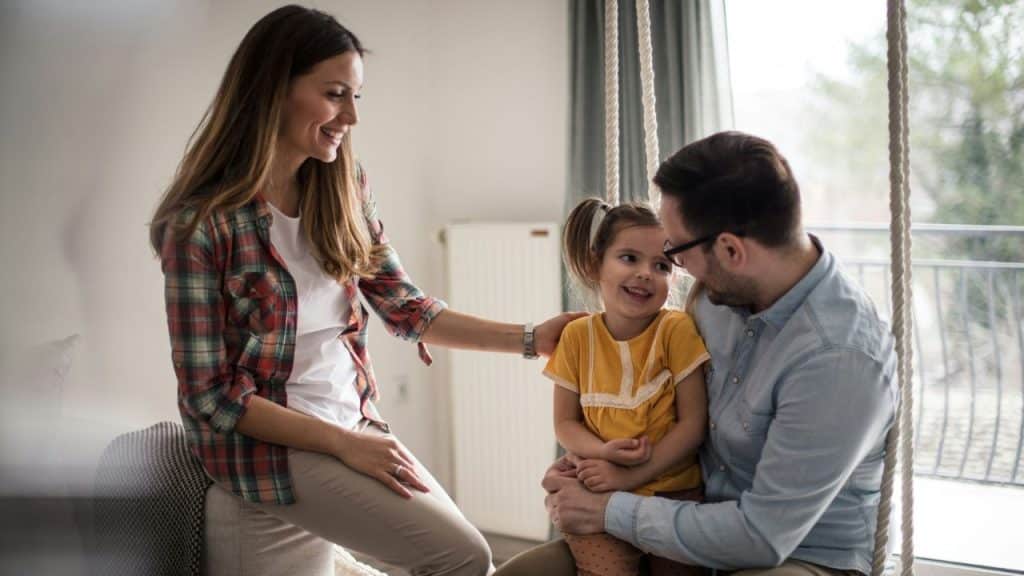 A father sits with his young daughter on an indoor swing, while the mother stands nearby, smiling.