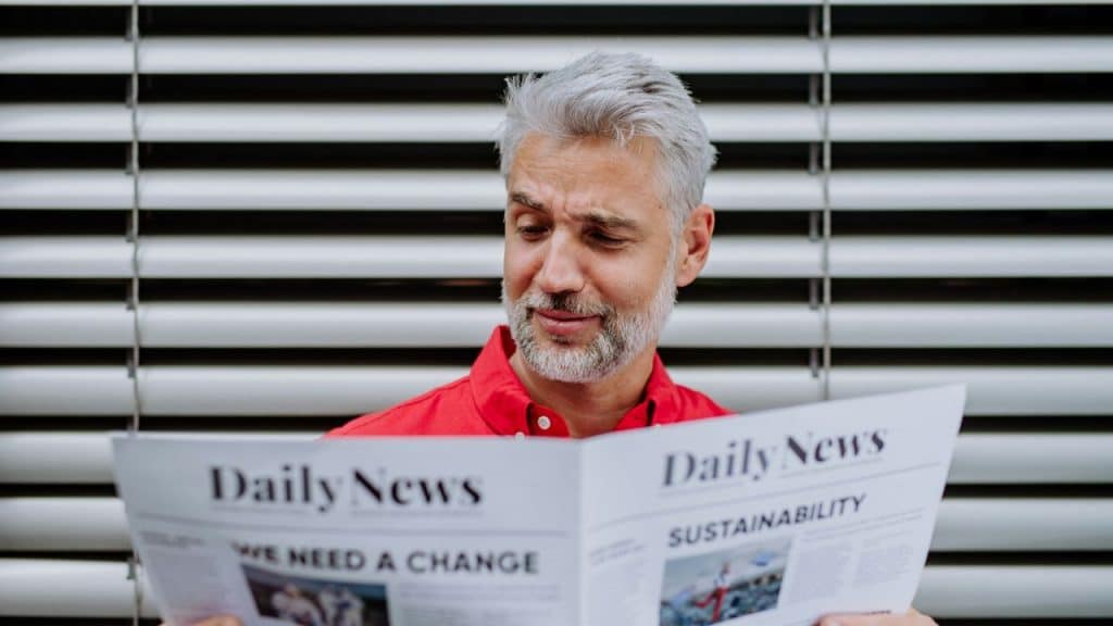 A gray-haired man in a red shirt reads a newspaper titled "Daily News" in front of horizontal blinds.