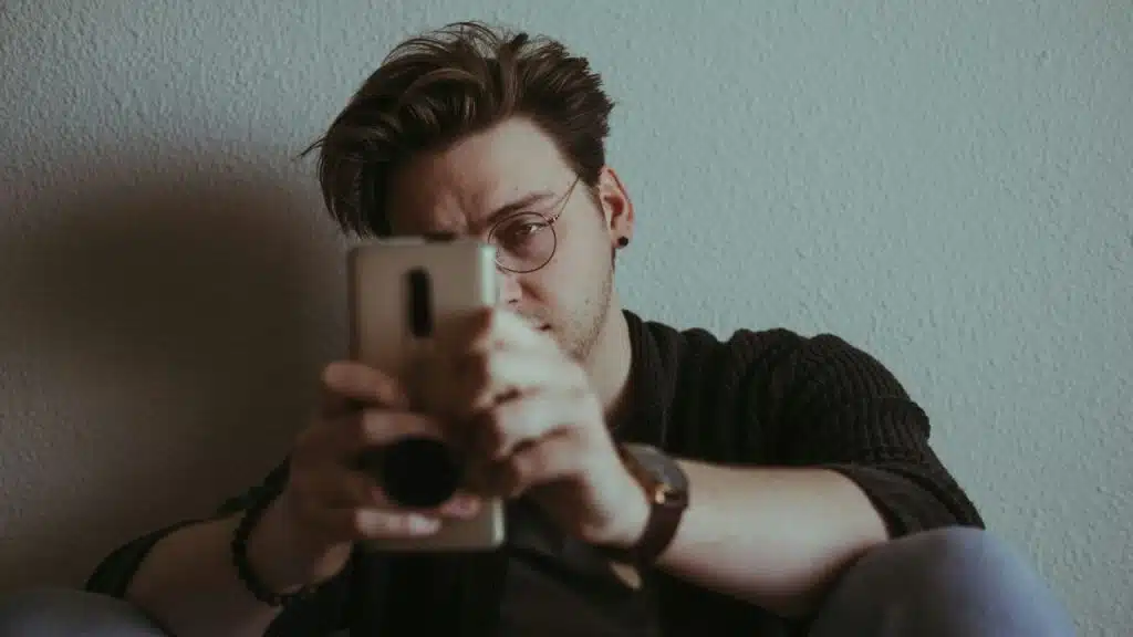 Man in a dark sweater and glasses holding a smartphone toward the camera.